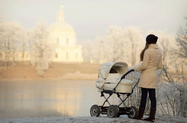 Mujer joven con abrigo blanco de pie cerca de cochecito de bebé blanco. Parque de invierno soleado, árboles helados y lago helado en el fondo — Foto de Stock