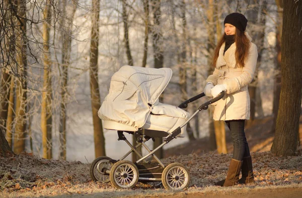 Sunny portrait of young woman in white coat standing near baby stroller in winter park with frosty trees — Stock Photo, Image