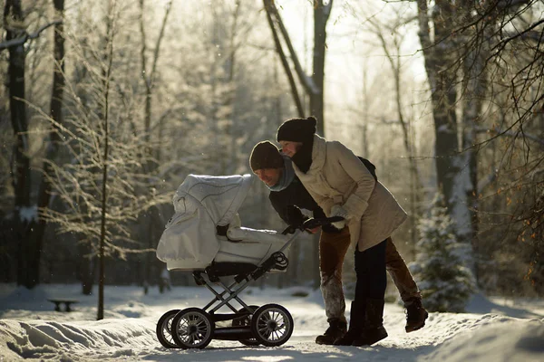 Молодые родители наклоняются над детской коляской, смотрят на ребенка. Солнечный зимний парк, ледяные деревья, забота о детях и любовь — стоковое фото