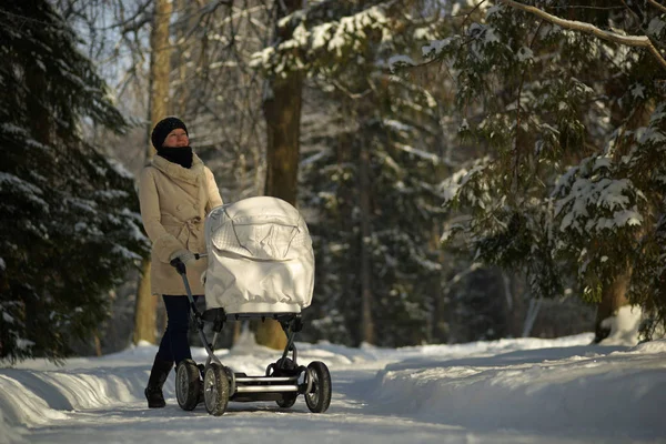 Молодая женщина в белом халате ходит с белой детской коляской в солнечном зимнем парке со снежными елками. Уход за детьми, любовь — стоковое фото