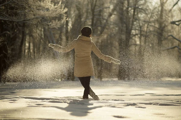Молодая женщина в белом халате и черной шляпе гуляет в солнечном снежном лесу на рассвете зимой, морозной солнечной зимой — стоковое фото