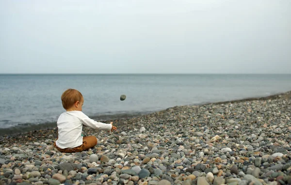 Menino sentado na praia de pedra e brincando com pedras. Vista por trás. Mar e céu nublado no fundo — Fotografia de Stock