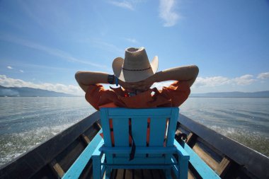 Young man in hat sitting on chair in boat with hands behind head and enjoying life, blue sky and clean water of lake clipart