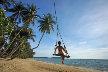 Young woman in bikini on rope swing on tropical sandy beach. Background of palm trees, blue sky and sea. View from back