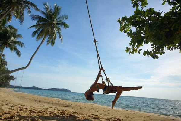 Elegant young woman swinging on rope swing on tropical sandy beach. Slim girl bending back. Palm trees, blue sky and sea
