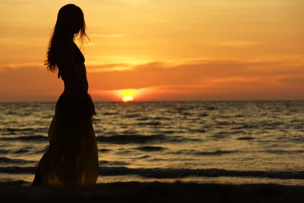 Silhouette noire de femme aux cheveux longs, en jupe longue debout sur la plage au coucher du soleil et regardant la mer — Photo