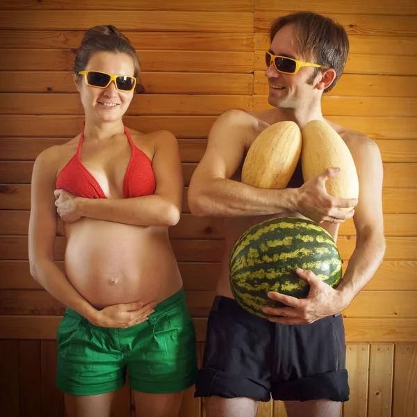 Divertido retrato de pareja. Mujer embarazada sosteniendo vientre y pecho, hombre sosteniendo watemelon en lugar de vientre y melones — Foto de Stock