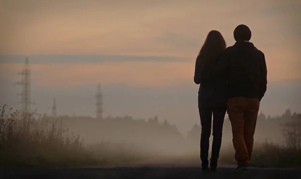 Silhueta preta de casal andando ao longo da rua enevoada no nevoeiro após o pôr do sol. Vista por trás. Homem segurando mulher — Fotografia de Stock