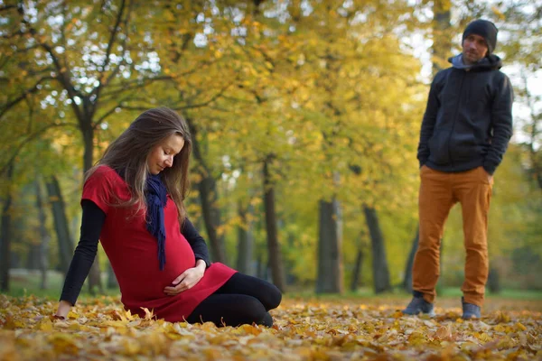 Joven mujer embarazada atractiva en vestido rojo sentado en hojas caídas en el parque. Hombre mirando a la mujer. 9 meses de embarazo — Foto de Stock