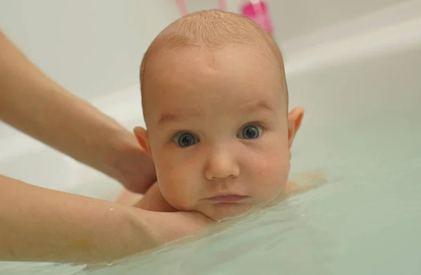 Evde küvette banyo yapan büyük gözlü, gülümseyen küçük bebek, suyun içinde bebeği tutan anne elleri. Yüzen çocuk