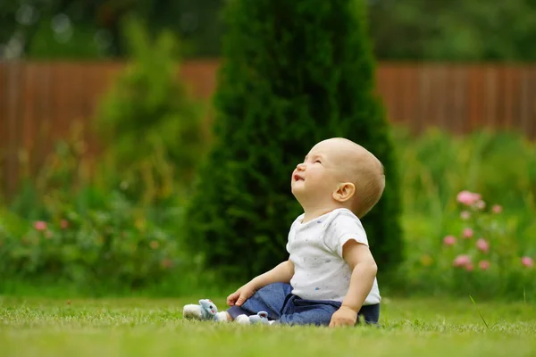Маленька мила усміхнена дитина в джинсах і білій сорочці сидить на траві в саду і дивиться вгору. Яскраво-зелений фон — стокове фото