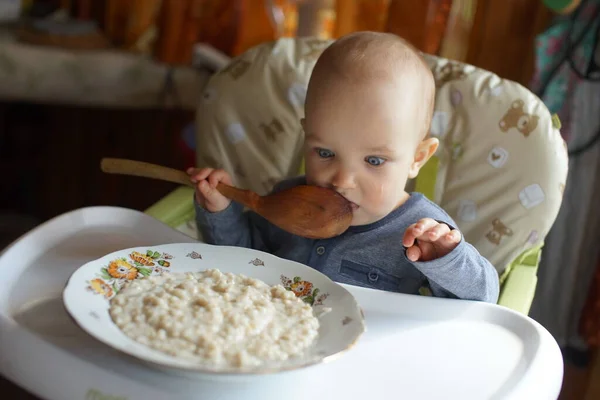 Bébé en chaise haute avec une énorme cuillère en bois, grande assiette avec du porridge devant, bébé regardant la plaque avec de gros yeux — Photo