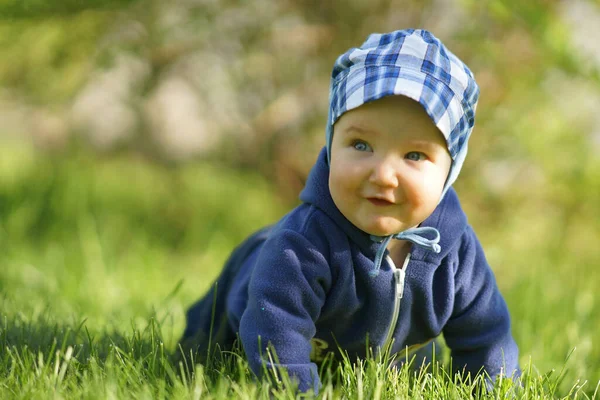 Маленька кумедна усміхнена дитина в блакитному одязі і кепка, що сидить на траві в сонячний літній день, яскраво-зелений фон — стокове фото