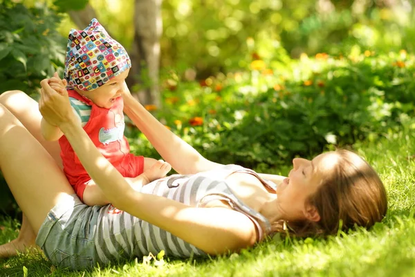 Giovane donna con piccolo bambino sdraiato sull'erba verde nella soleggiata giornata estiva. Ridere bambino seduto sulla madre sorridente — Foto Stock