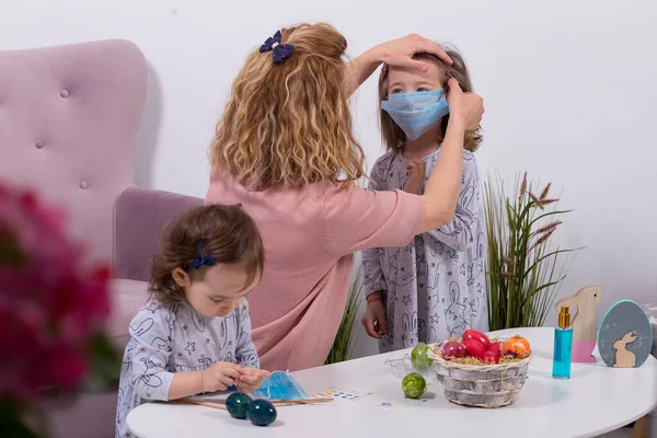 Anne ve kızları yumurta boyuyor. Mutlu aile Paskalya için hazırlanıyor. Annesi Coronavir 'e karşı kızının maskesini takıyor. Evde karantina.