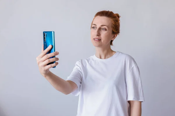 Kadın gülümsüyor, beyaz tişört giyiyor, beyaz arka planda izole edilmiş bir cep telefonu kullanıyor.