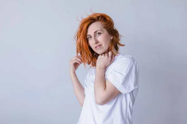 白いカジュアルなTシャツを着て カメラで楽しそうに見て 長い赤い髪をした快適な白人女性の横の肖像画 ロイヤリティフリーのストック写真