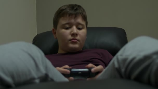 若い男の子が座って アームチェアでリラックスし ビデオ ゲームをプレイします ゲーム コント ローラーを使用し — ストック動画