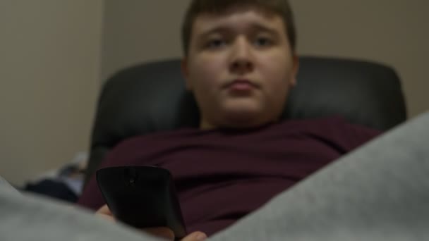 若い男の子に座って肘掛け椅子でリラックスとテレビを見てテレビを保持している彼の手でリモート — ストック動画