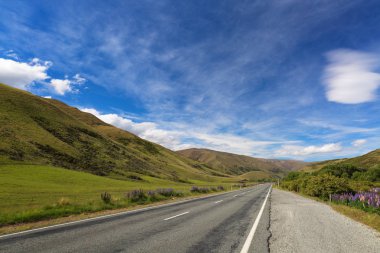 A straight country road between green hills in New Zealand. Bright blue sky with white clouds, sunny day clipart