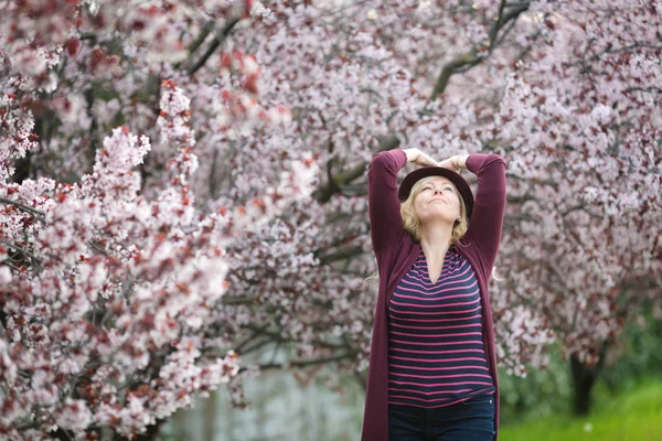 Çiçek açması tree yakınındaki mor fedora şapkalı uzun saçlı beyaz sarışın kadın gökyüzünde mutlu arıyor