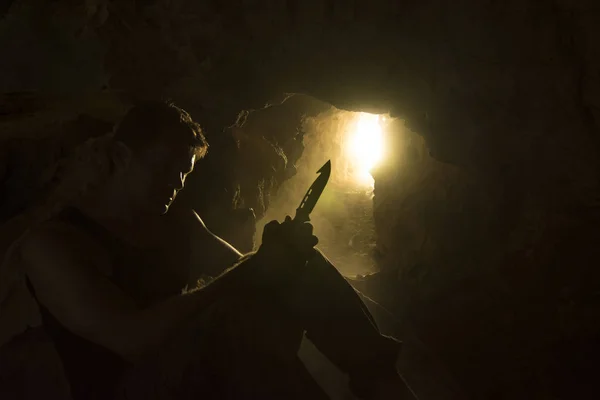 Survival man with knife backlit in dusty cave