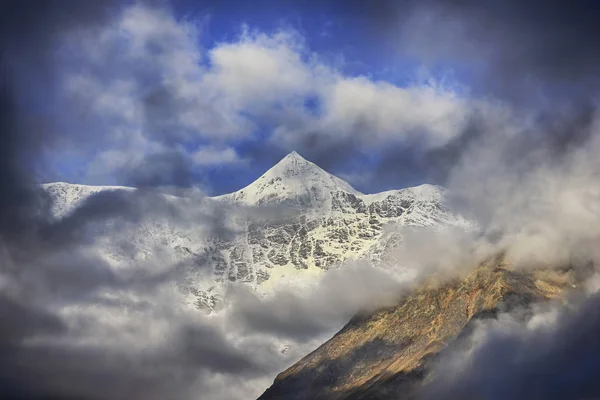 Vista sulla cima alta montagna ghiacciata illuminata dal sole attraverso le nuvole . — Foto Stock