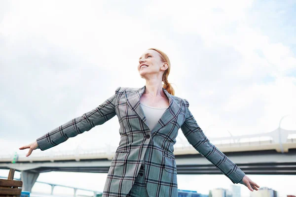 Low angle view of cheerful middle-aged businesswoman standing on upper deck of ship with outstretched arms and enjoying breathtaking view, portrait shot
