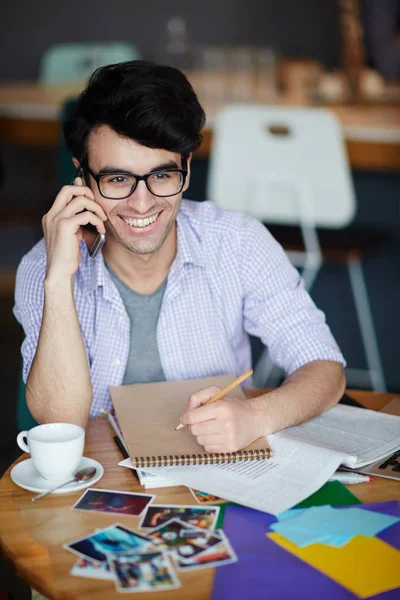 カジュアルな服装と笑顔と電話で呼び出している間ノートを作ってテーブルに座ってメガネを身に着けている創造的な若者の肖像 — ストック写真