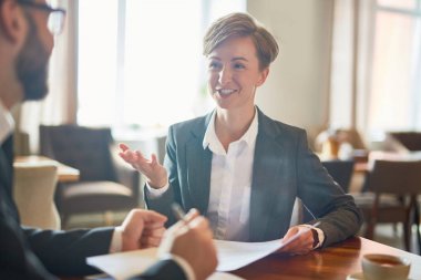 Modern rahat kafede otururken iş ortağı ile müzakereleri yürüten resmi kıyafeti neşeli güzel girişimci