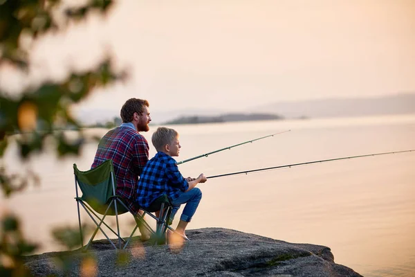 両方身に着けている格子縞のシャツ 木の後ろから撮影の風景と水の父と岩の穏やかな湖の棒で釣りに一緒に座って息子の側ビュー肖像画 — ストック写真