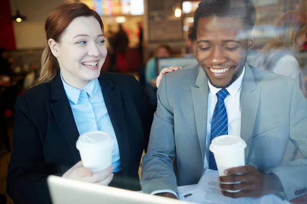 Two business people meeting in coffee shop after work: Young woman and African-American businessman looking at computer screen, laughing and drinking coffee, shot behind glass window