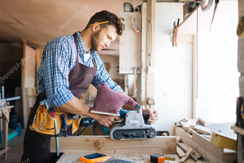 Young bearded carpenter in apron sanding plank in workshop, workbench covered with shavings