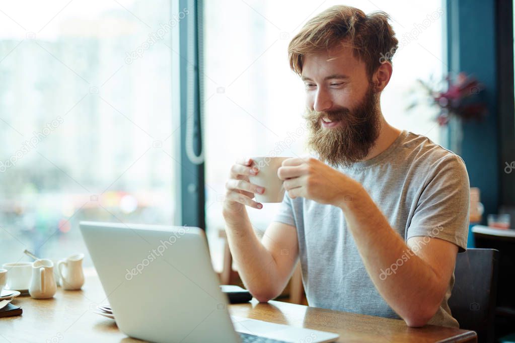 Cheerful bearded businessman enjoying delicious coffee while sitting in lovely small cafe and working on laptop, waist-up portrait