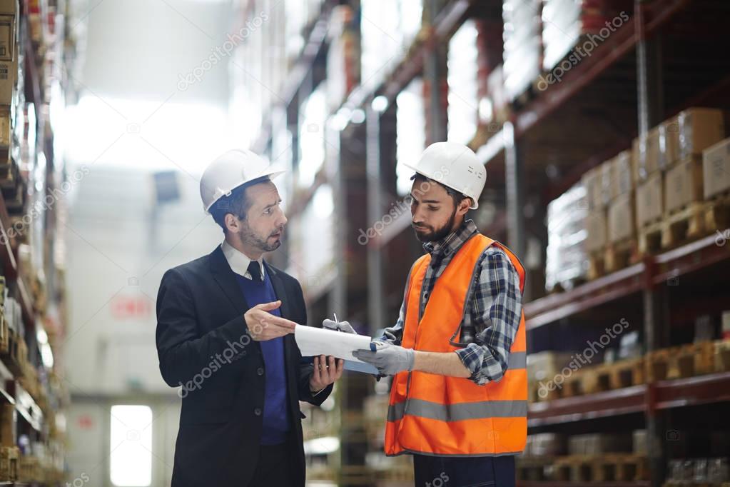 Portrait of warehouse manager wearing business suit talking to loader showing order list in aisle between tall shelves with packed goods