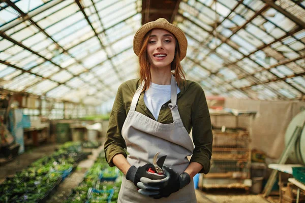 植物での作業を楽しんでいる麦わら帽子をかぶって笑顔の美しい女性の肖像画花保育園でカメラ目線に立って — ストック写真
