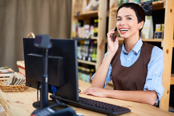 Waist-up portrait of attractive middle-aged store owner looking at camera with wide smile while having telephone conversation with supplier of organic products