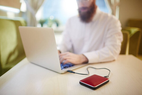 Занят молодой предприниматель с густой бородой, работающий над перспективным проектом с помощью ноутбука, подключенного к электроэнергетическому банку, сидя в милом кафе
