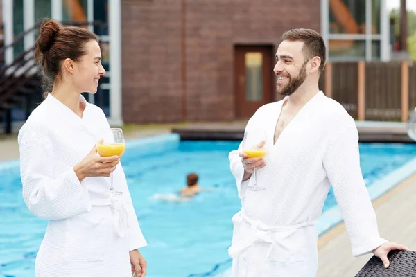 スイミング プールでオレンジ ジュースを持つ白いバスローブで幸せな若いカップル — ストック写真