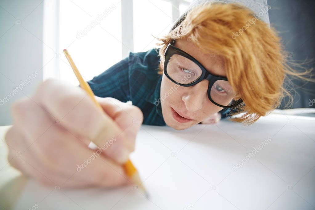 Portrait of young creative red haired man wearing beanie hat and glasses drawing with pencil leaning close to desk and looking focused