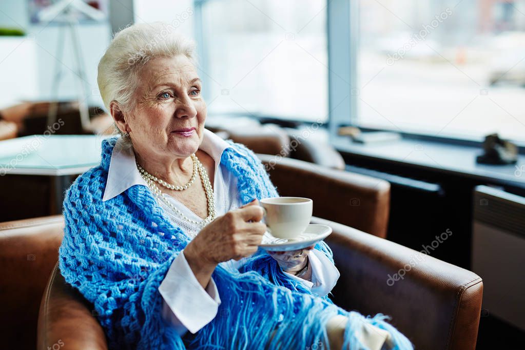 Portrait of elegant-looking elderly woman with cornflower blue knitted shawl on shoulders holding cup of delicious coffee in hands, sitting in comfortable armchair and enjoying picturesque view from window 