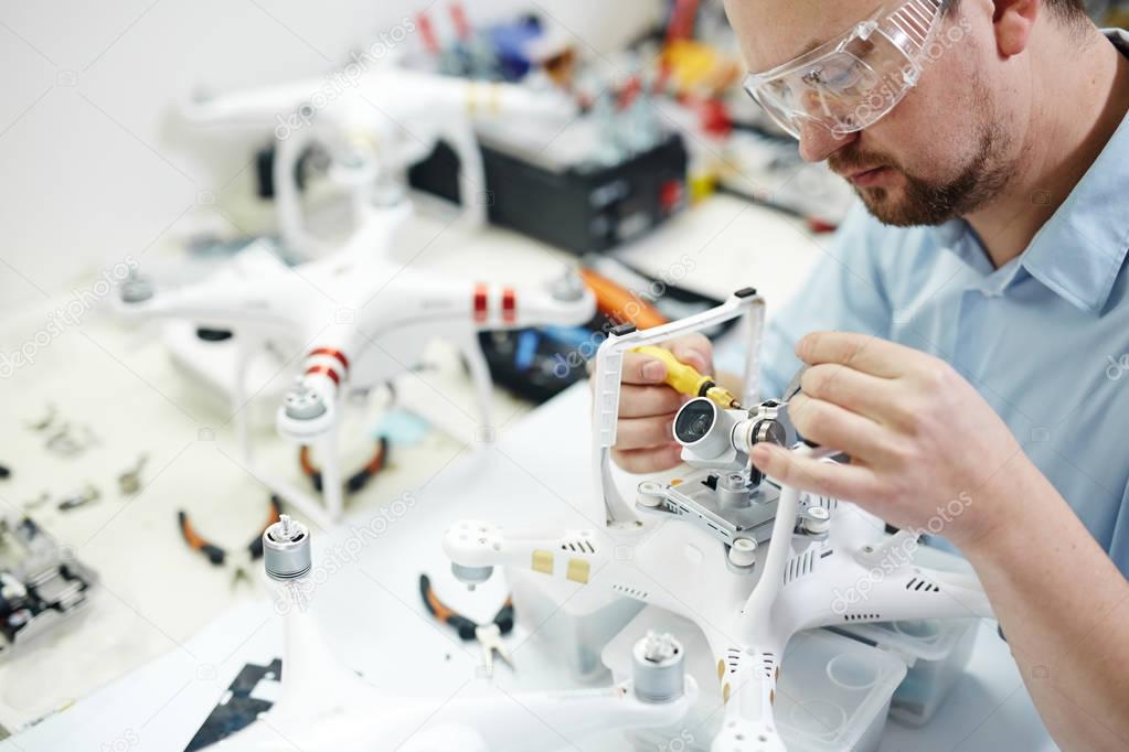 Man in protective glasses working on assembling new spy system connecting  quadcopter drone and action camera on table  with different tools in modern workshop