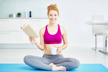 Yoga yaparken uygun kırmızı saçlı kadın portresi egzersizleri evde: lotus pozisyonda bacaklar ile oturan geçti iken gülümseme ile kitap okuma ve mutlu bir şekilde gülümseyerek kameraya, seyir