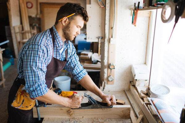 Skillful bearded woodworker in checked shirt smoothing plank with jointer plane, shavings scattered on table