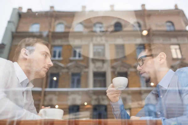 两个商人坐在咖啡馆对面的餐桌边上 在会议上讨论交易 从玻璃窗后面开枪 — 图库照片