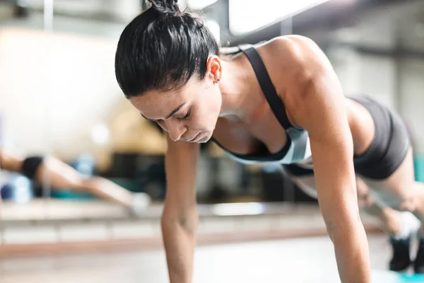 在现代健身房锻炼时用力做板运动的坚强的肌肉妇女的画像在镜子旁边 — 图库照片