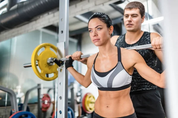 私人教练的肖像帮助美丽的肌肉妇女穿着运动服做举重运动在现代健身房中肩扛着大的重杠 — 图库照片