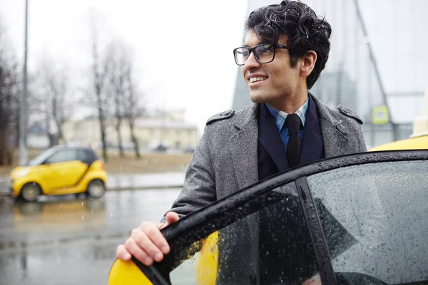 年轻英俊的中东商人的画像在下雨的秋天街道乘坐出租汽车 微笑并且看着一边打开车门 — 图库照片