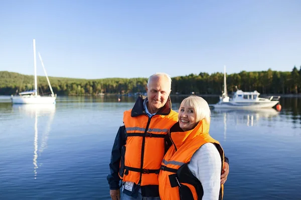 Happy seniors in life-vests standing by lake on summer day