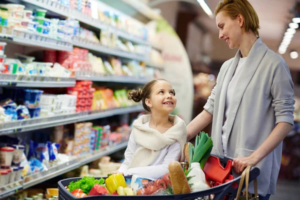 小さな女の子 笑顔と食品のカートを押しながらに渡ってお互いを見てと食料品店のショッピングに幸せな女性の肖像画 — ストック写真